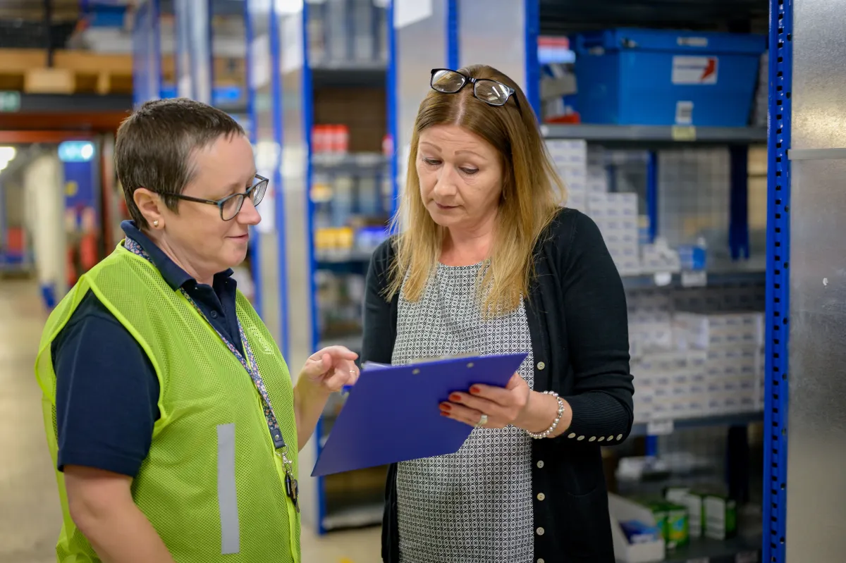 Two staff members reviewing a clipboard beside warehouse shelving.