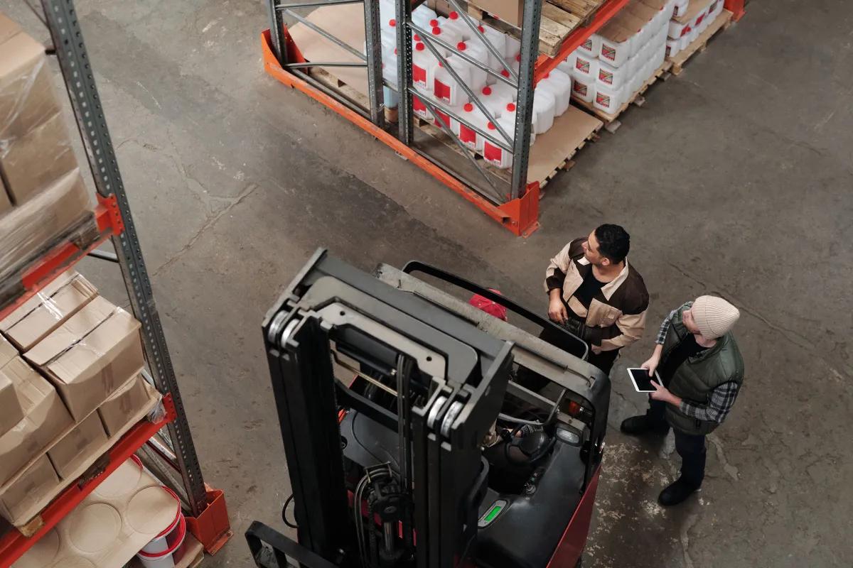 Two warehouse workers discussing a load beside a forklift and pallet racking, seen from above.